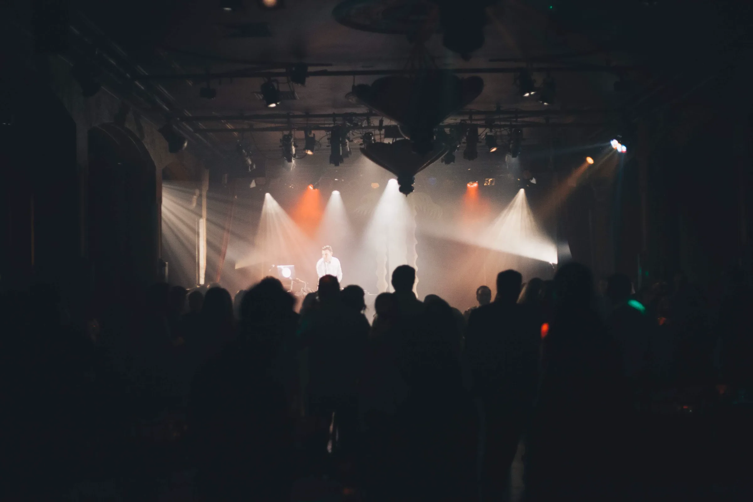 Wide stage with white and amber light beams cutting through haze over a full crowd